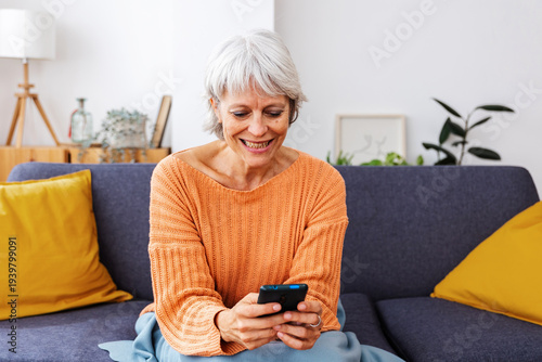 Schilderij op canvas Happy senior woman enjoying winter day at home while entertaining using mobile phone, reading some news or watching social media content on app