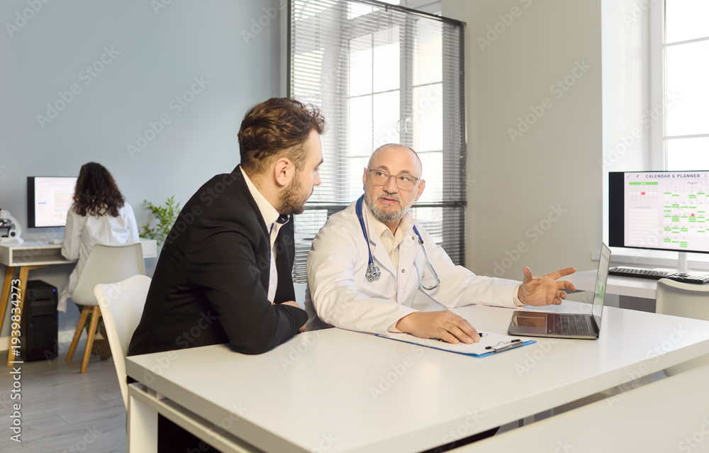 Fototapeta premium Doctor patient consultation in medical clinic office. Senior physician gives diagnosis and advice using a laptop to explain results to the patient. Concept: trust and professional care.