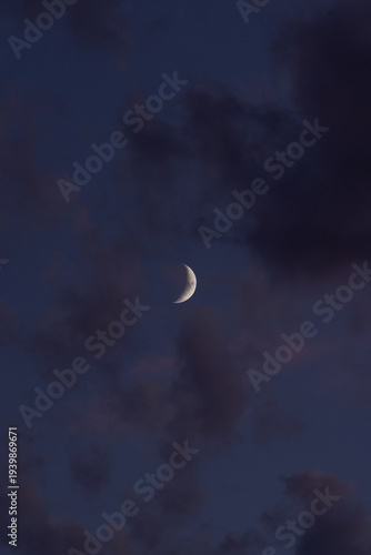 Crescent moon glows softly through deep twilight clouds. Dark, textured clouds drift around the serene lunar curve. Night sky feels calm, quiet, and mysterious. Ideal for tranquil scenes, romance