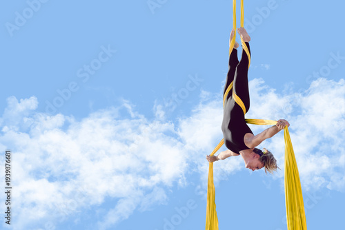 Young athletic woman hangs upside down forming a cross shape with open arms while suspended by aerial silk fabric, training outdoors to improve strength, balance, flexibility, and body control