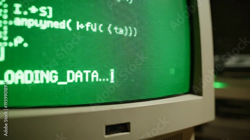 Close view of a vintage computer monitor showing loading data text and programming code, set in a dim workspace with green glow, conveying retro tech focus