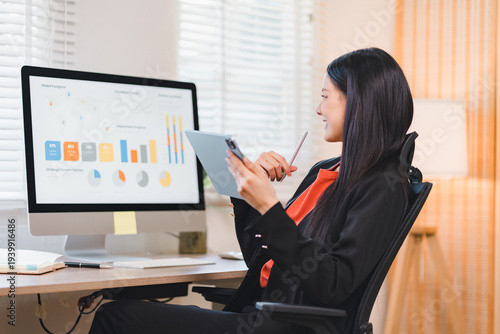 Happy Asian female using tablet technology in business office to view finance chart analysis on computer monitor with cheerful smile and success