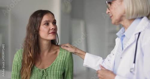 Closeup of smiling female doctor talks with older adult woman patient during medical consultation in clinic, touching her shoulder, explain procedure, share good health news, supporting and reassuring