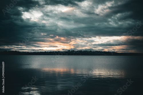 Fotografia Dramatic sunset over a calm lake with a distant tree line and moody clouds