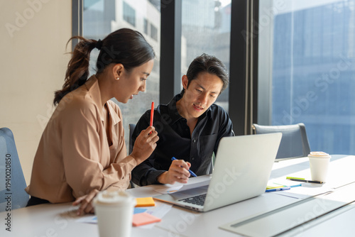 Asian business woman and man using laptop working and discussing project details on documents together in modern office. Two coworkers brainstorming and meeting table in workspace
