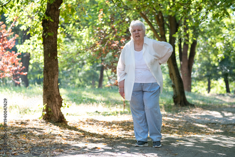 Fototapeta premium Senior woman feels pain while walking in park in summer. Elderly lady touching back by hands, having backache, muscle, hip pain outdoors. Old people suffering from waist pain.