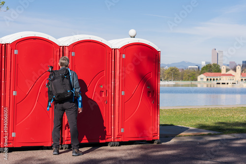 A man with a black backpack standing in front of a row of red portable toilets at a sunny lakeside park.