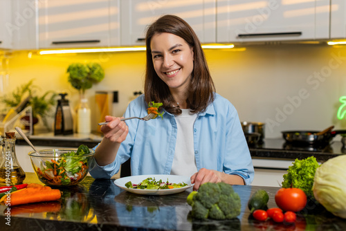 Young woman eating salad in home kitchen smiling while choosing healthy food for diet weight control. Adult girl enjoying lunch break with bowl using fork feeling satisfied calm indoors for wellness.