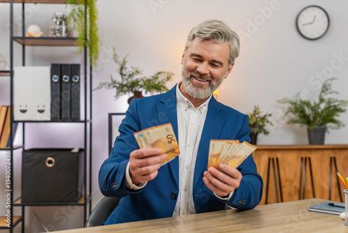 Fotografie Middle-aged businessman at home office waves euro bills counting fresh salary income feeling triumphant