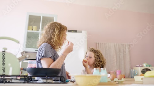 Cute curly haired daughter and her mother tasting fresh homemade food while cooking breakfast together. Happy family enjoying a sweet moment
