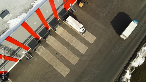 High angle drone shot slowly descending above warehouse loading docks as a cargo truck reverses into a distribution center bay and driver walks toward the building entrance.