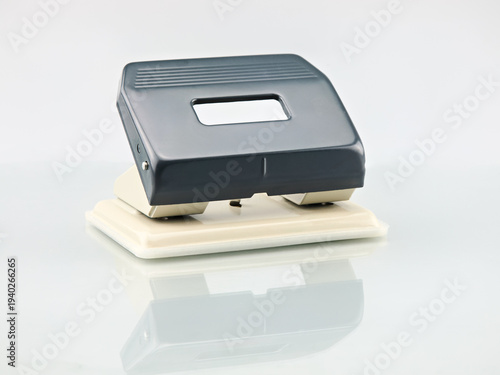 Konstfotografi A sleek dark grey and beige two-hole puncher sits on a reflective white surface, ready for office use
