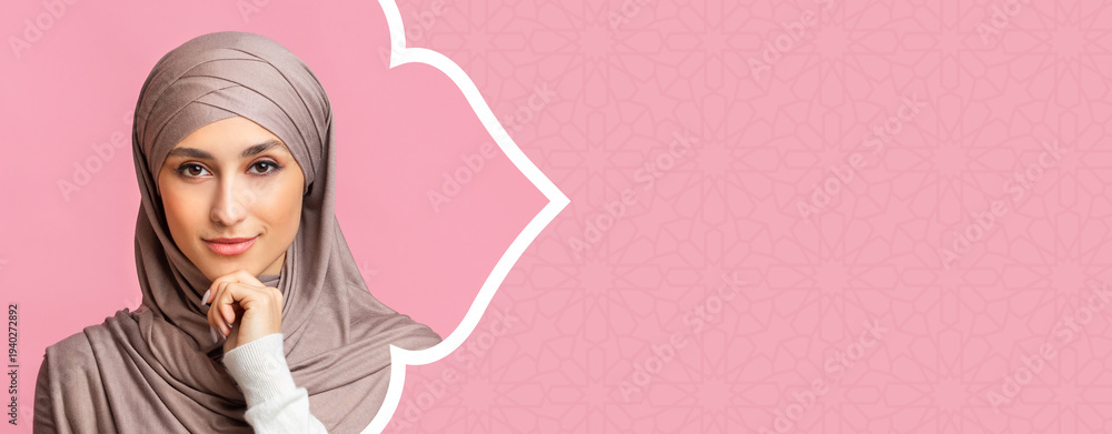 Naklejka premium A young woman is shown in a headscarf. She has a gentle look on her face and rests her chin on her hand. The background is pastel pink with a subtle design.
