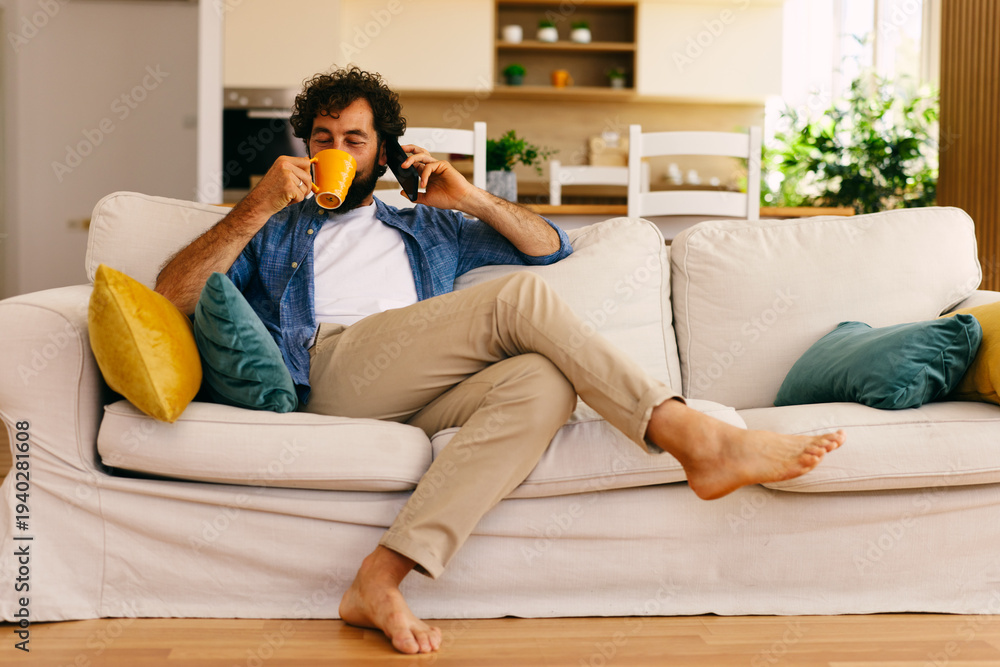 Fototapeta premium Man drinking coffee while talking on smartphone at home