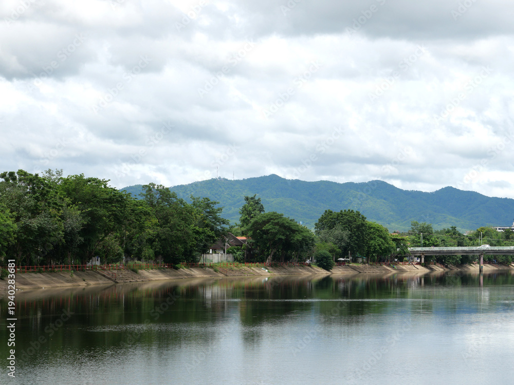 Fototapeta premium Calm river reflecting green trees and a bridge under a cloudy sky.
