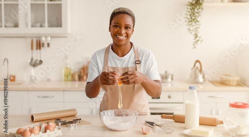 Fotografi A woman stands in a well-lit kitchen, cracking an egg into a bowl while smiling