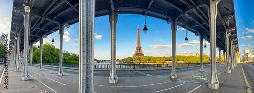 A clear blue sky frames the iconic Eiffel Tower, observed from under the Bir-Hakem Bridge. The scene captures the beauty of Paris along the Seine River on a tranquil afternoon.