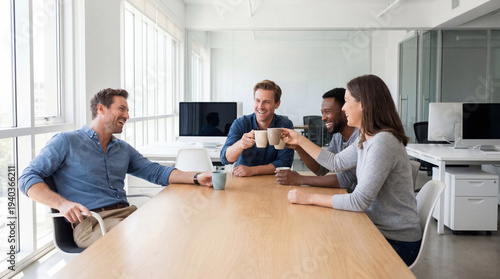 Four colleagues toasting with coffee mugs and smiling in spacious bright corporate office with glass walls