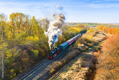 defaultAn old black steam locomotive pulls a passenger train through a magical autumn forest. Aerial view.