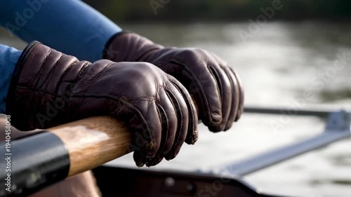 A close-up view showing a rower's gloved hands firmly gripping an oar handle, poised above the water, highlighting the focused determination and physical effort in the sport of rowing