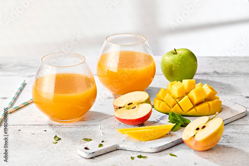 Apple and Mango Juice in Short Round Tumblers with Fresh Apples and Mango on White Wooden Surface