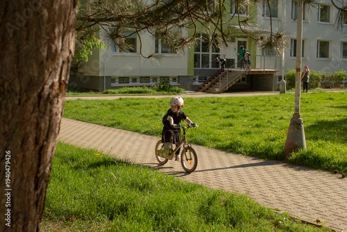 Child on bicycle on paved path, helmeted youngster riding near grassy apartment courtyard, warm sunlight casting long shadows. Spring