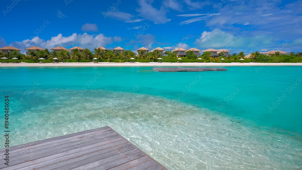 Fototapeta premium View from a wooden deck looking out over crystal clear turquoise water towards a row of beach villas on a tropical island
