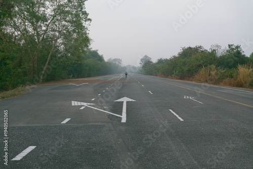 A lone cyclist rides along an empty rural road with directional arrow markings disappearing into a misty countryside landscape.