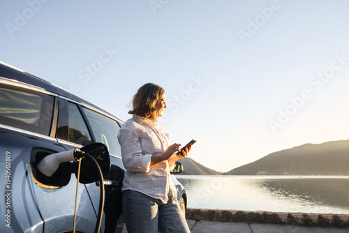 Wallpaper Mural Woman with smartphone next to charging electric car on the seacost during summer vacation Torontodigital.ca