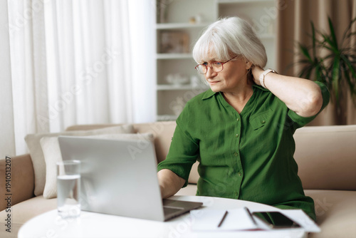 Senior woman touching neck with discomfort while sitting at laptop at home. Mature caucasian female experiencing neck pain from computer use, highlighting health and ergonomic issues