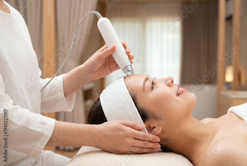 笑顔で美容クリニックのフェイシャル治療を受ける若いアジア人女性、エステティシャンによるスキンケアと美顔器の施術、リラックスしたスパの背景