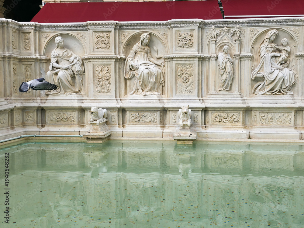 Fototapeta premium The ornate marble reliefs of Fonte Gaia fountain with water reflections and a flying pigeon in Piazza del Campo, Siena, Tuscany, Italy