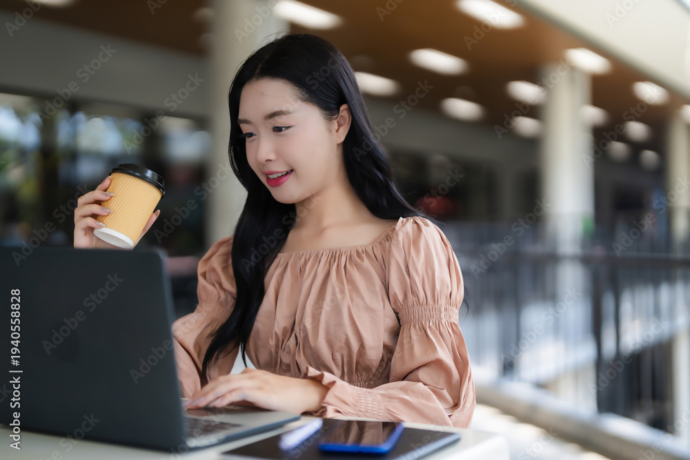 Fototapeta premium Asian young adult woman freelancer works laptop computer holding coffee cup modern cafe casual professional remote work business education student online