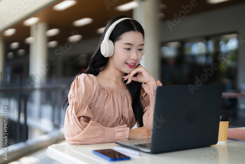 Asian adult woman uses laptop headphones learn work study engage online content education business communication modern cafe coworking space remote environment