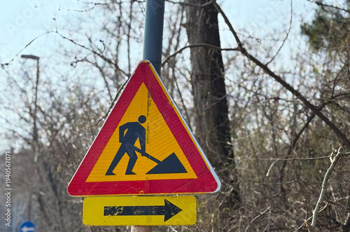 Roadwork warning sign with arrow direction placed near forest roadside. Traffic regulation, infrastructure maintenance, driver attention, safety awareness and transportation management concept.