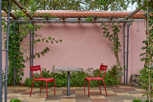 A serene outdoor patio scene featuring two vibrant red metal chairs facing each other across a small wooden table, set against a pastel pink wall. The courtyard is framed by a rustic wooden pergola w