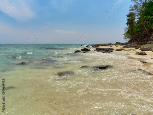 On the sandy shore, sea waves are foaming, there are large dark stones, a blue sky with light clouds, and the sea is slightly rough