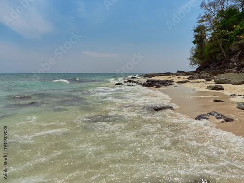 On the sandy shore, sea waves are foaming, there are large dark stones, a blue sky with light clouds, and the sea is slightly rough