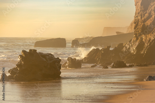 Golden sunset light illuminates rocky shoreline with ocean waves and misty sea spray, creating a peaceful coastal landscape with warm tones, rugged cliffs and natural seaside atmosphere