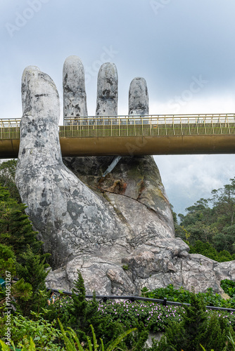 Golden bridge structure appears held by large stone hands amidst green mountain landscape in huyn hoa vang, da nng
