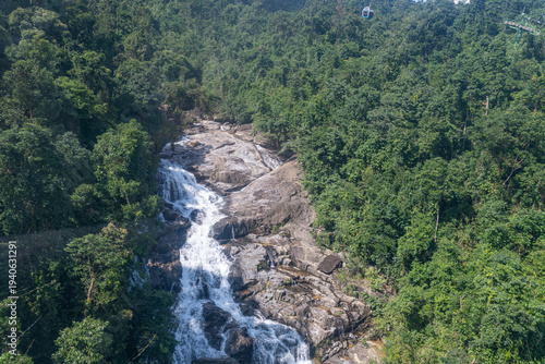 Wild waterfall stream cascading among lush green jungle trees with distant cable car gondola