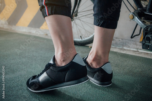 Close up of female cyclist leg standing with road racing bike.Cycling Shoe and a bicycle wheel at a road.Female leg biker in Black cycling sneaker shoe beside bicycle.