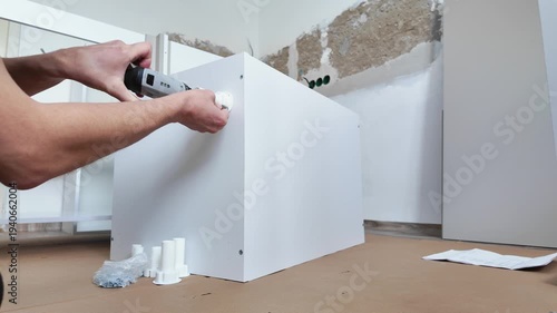 Wide Angle Contractor Securing Cabinet To Wall Caucasian Installer Using Power Driver To Fasten Brackets And Anchors, Reinforced Alignment, Protective Cardboard On Floor, Hardware Pile Close By