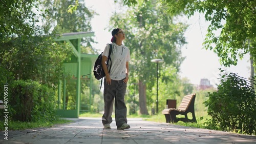 Strolling man under leafy shade, Person leisurely walking past greenery with backpack on warm afternoon, Individual taking slow walk in park with trees and backpack on sunny summer day