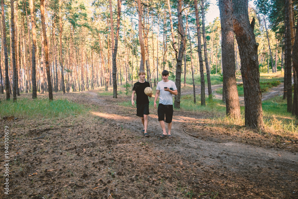 Fototapeta premium Two Teenage Friends Walking through a Sunny Pine Forest with a Soccer Ball and Smartphone during Golden Hour