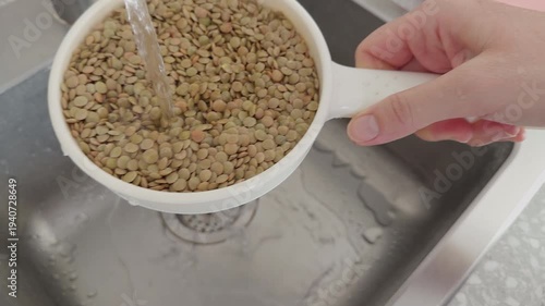 A person rinsing dry green lentils in a white plastic colander under a stream of fresh tap water in a kitchen sink. Close up.