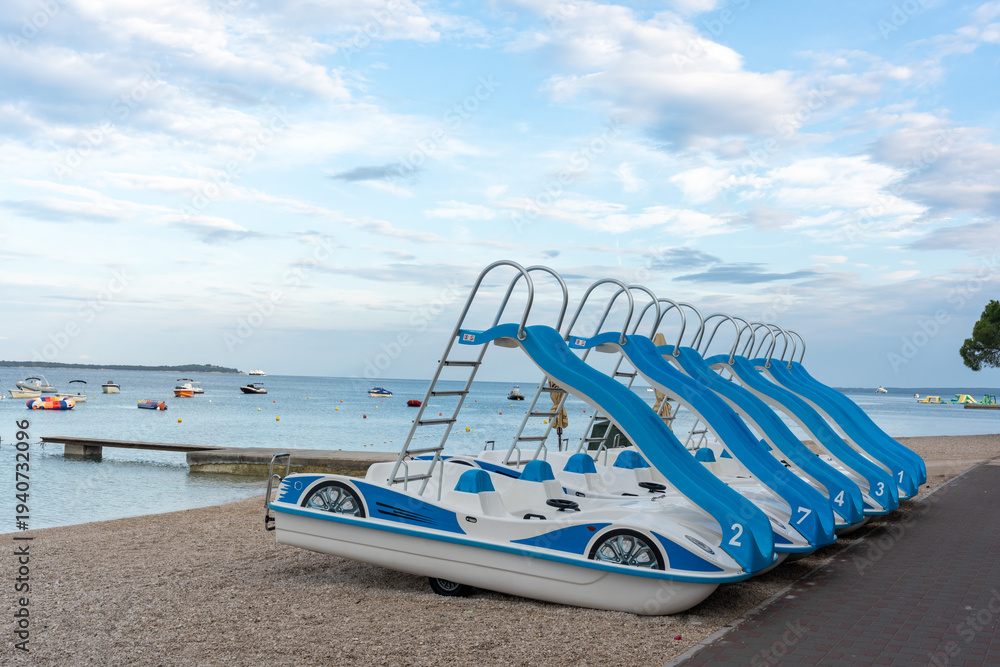 Obraz premium Row of blue and white pedal boats with water slides lined up on pebble beach ready for summer rental with calm sea. Beach water entertainment, family fun and active seaside holiday.