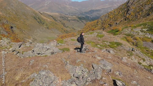 Lone figure stands atop rugged terrain beside a turquoise lake. The person gazes out at the vast, sunlit valley beyond. Sunlight glints off the water and rocks in golden hues