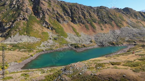 Aerial view reveals a turquoise lake nestled in rugged mountains. Snow-capped peaks frame the serene, glacial-fed waters. Golden grasses surround the valley, hinting at autumn's approach