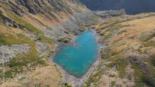 Aerial view reveals a turquoise lake nestled in rugged mountains. Snow-capped peaks frame the serene, glacial-fed waters. Golden grasses surround the valley, hinting at autumn's approach